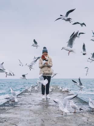 Happy young tourist woman feeds seagulls on the sea. Pretty female wearing coat, scarf and watching flying seagulls by the sea in the sky. People, travel, environment, nature concept.