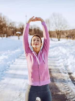 Three quarter length of beautiful Caucasian brunette in sportswear standing on country road and stretching arms before running. Wintertime.