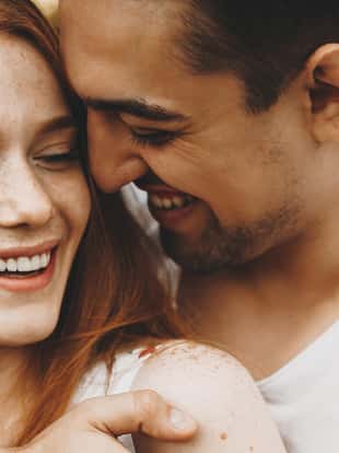Close up portrait of a charming young couple laughing while man is embracing her girlfriend against sunset outdoor in park.