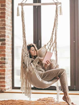 Beautiful young woman, which is wearing cashmere nightwear clothes, reading novel on rocking chair infront of big window in scandinavian cabin one lazy weekend morning