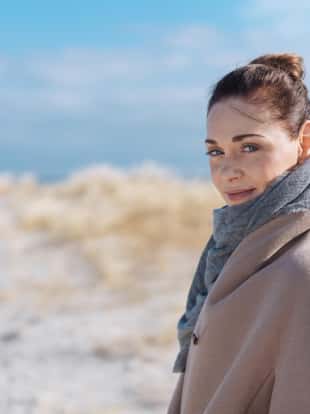 Attractive woman in coat and scarf standing on a sunny sandy winter beach turning to look thoughtfully at the camera with copy space
