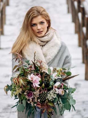 Beautiful woman stands on the wooden bridge in snowy forest in grey coat with bouquet in hand