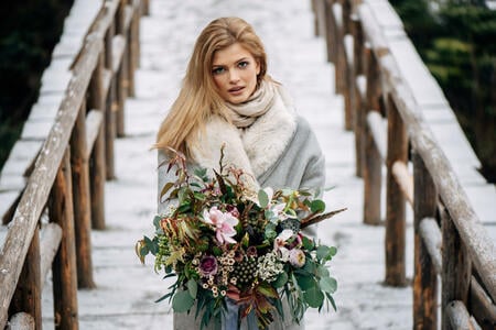 Beautiful woman stands on the wooden bridge in snowy forest in grey coat with bouquet in hand
