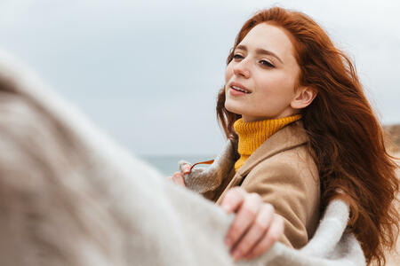 Beautiful young redheaded woman wearing coat, covered in blanket walking at the beach