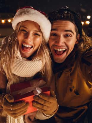 Cheerful young couple dressed in winter clothing holding gift boxes sitting outdoors, taking a selfie, snowfall