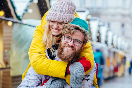 Couple in warm bright outwear, man giving piggyback to woman in street, outdoor. Handsome beard hipster man in eyeglasses and his girlfriend having fun in winter city. Cose up, portrait. High emotions
