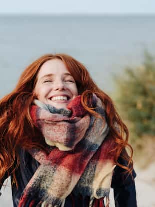 Joyful happy young redhead woman wearing a warm winter scarf looking up into the air with a beaming smile as she strolls on a sandy beach in the dunes