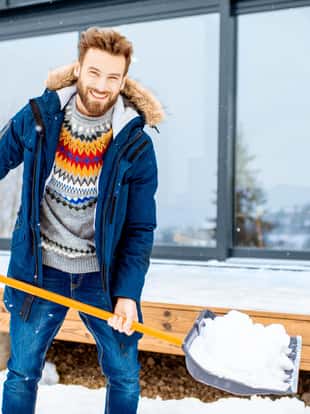 Handsome man in winter clothes cleaning snow with a shovel near the modern house in the mountains