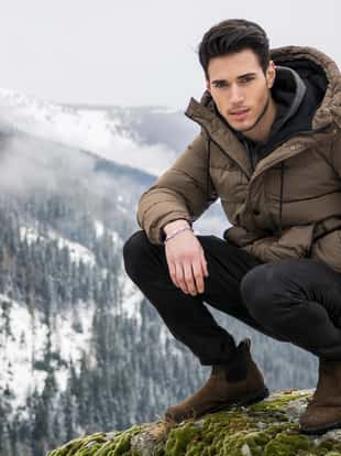 Handsome man in outerwear sitting while looking at camera. Snowy landscape on background