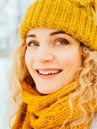 Outdoor close up portrait of blond curly smiling hipster girl posing on street, looking at camera, smiling at snowy park. Model wearing white sweater, yelow winter hat, sarf, gloves. City lifestyle
