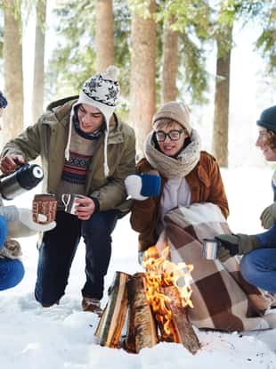Full length portrait of four young people camping in winter forest sitting in circle round fire and drinking hot cocoa