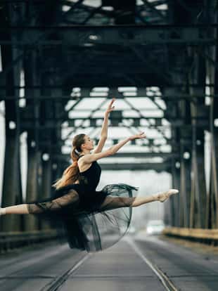 Ballerina jumping in twine pose in a black transparent dress on the road and rails against the background of metal supports.