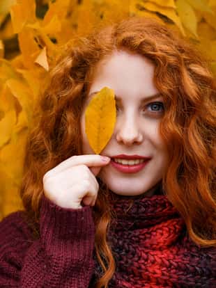 cute red-haired girl on the background of an autumn tree with a leaf in her hand smiles and grimaces