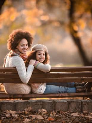 Happy black girl and her mother relaxing on a bench in autumn park and looking at camera.