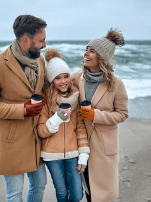 A beautiful married couple and their charming daughter walk on the seashore and drink coffee, dressed in warm clothes.