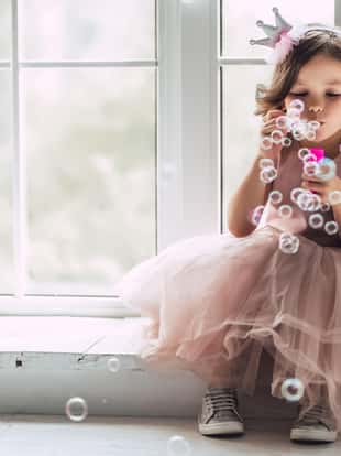 Little cute girl in beautiful dress is sitting near the window at home and blowing soap bubbles.
