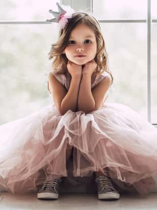 Little cute girl in beautiful dress is sitting near the window at home.