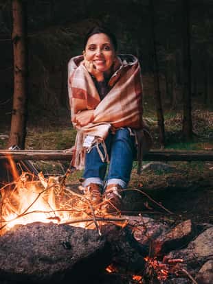 Pretty smiling young woman sits near the campfire in twilight autumn forest