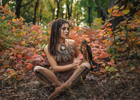 Aggressive-sexual wild girl, wanders in the jungle with a tamed bird. Princess warrior in a brown dress and a necklace of feathers. Background autumn landscape. Hairstyle dreadlocks. Artistic Photography