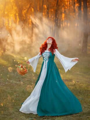 charming girl with bright red hair in national Bavarian green costume joyfully dances on green meadow near summer forest, magical sorceress collected fresh apples basket in rays of warm setting sun.