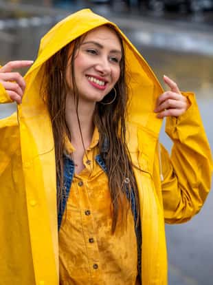 Happy young woman preparing for rain. She is wearing a yellow raincoat and she is putting the hood on so she can cover her head.