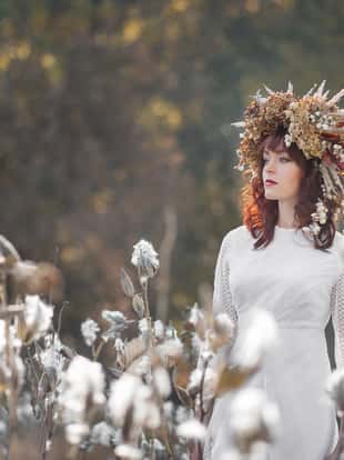 Young beautiful girl in a white vintage dress and wreath of dried flowers on the head in a autumn field. Mysterious fluffy seeds of a of opened Asclepias Syriaca pod (Milkweed, Silkweed).