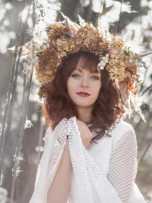 Young beautiful girl in a white vintage dress and wreath of dried flowers on the head in a autumn field. Mysterious fluffy seeds of a of opened Asclepias Syriaca pod (Milkweed, Silkweed).