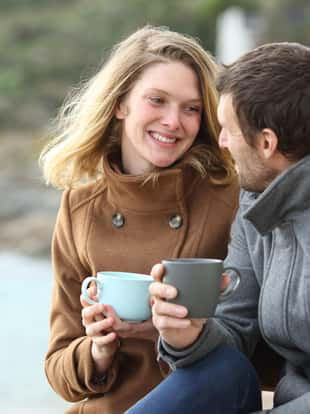 Happy couple of adults talking drinking coffee sitting outdoors in winter on the beach