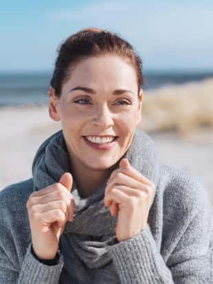 Attractive woman on a sunny beach in cold weather holding her grey scarf with her hands as she looks aside with a lovely friendly smile