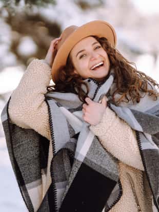 Beautiful woman standing among snowy trees and enjoying first snow. Young woman wearing hat, plaid scarf and coat. Happy winter time. Christmas.