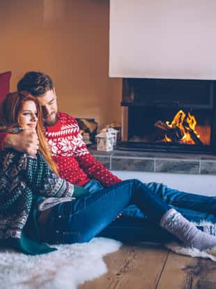 Couple on vacation at mountain cabin. Sitting on the floor on a fur by fireplace in cozy  living room on Christmas. Wearing festive knitted sweaters and socks. Austrian Alps.