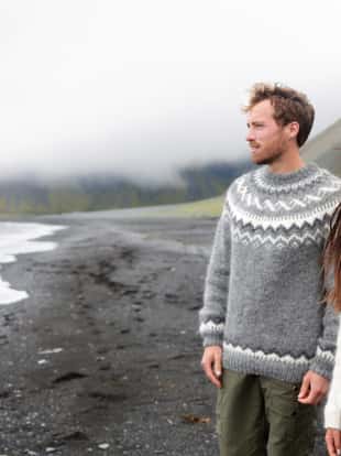 Iceland couple wearing Icelandic sweaters on black sand beach. Woman and man model in typical Icelandic sweater looking at ocean enjoying view of nature landscape at ocean sea.