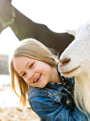 Pretty little blond girl in denim jacket with goat and horse on sunny winter day.