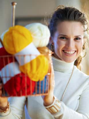 Portrait of smiling trendy female in white sweater and skirt showing basket with knitting yarn and needles at modern home in sunny winter day.
