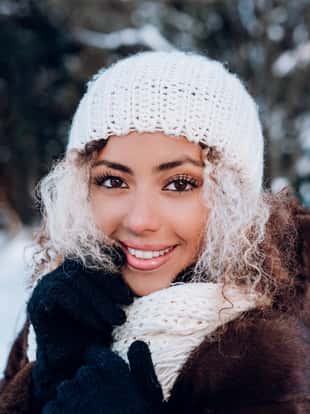Smiling young afro american woman touching face, wearing stylish white knitted winter hat and gloves in winter park.