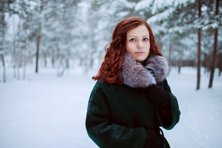 An attractive girl with red curly hair stands in the winter forest, snow falls, she is wrapped in a coat with a fur collar.