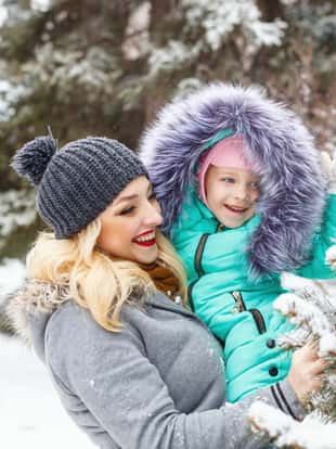 Mother holds daughter on hands. They stand next to the Christmas tree in snow. Family time. Winter walk in the park.