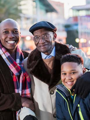 A multi-generation African-American family portrait. A mid adult man in his 30s standing with his 10 year old son and his 79 year old grandfather, who is the boy's great grandfather. They are in the city by the waterfront in winter or autumn, wearing coats and scarves.