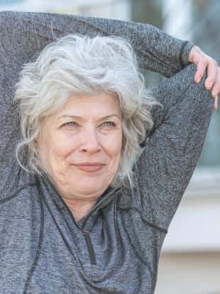 A confident senior woman with grey hair is attending a fitness class outdoors. She is stretching her arms and smiling. She is relaxed and excited to exercise.
