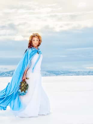 Beautiful Snow Queen Walking Across A Lake of Ice and Snow. She wears her Tiara, her icy blue velvet cape, a snowy white dress and holds a frozen bouquet of blue roses. She is looking at the camera. The wind is blowing her cape and hair as nature swirls all around her. Cold Beauty. Copy Space.