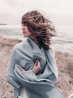 Attractive young woman standing on a windy cold beach wrapped in warm blanket with flower in her hand. Windy cold weather concept.