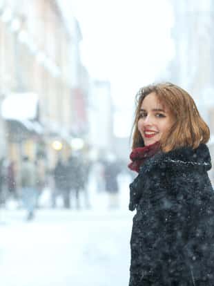 Emorional brunette girl with red lips wears coat and scarf walking at the city during snowfall. Empty space