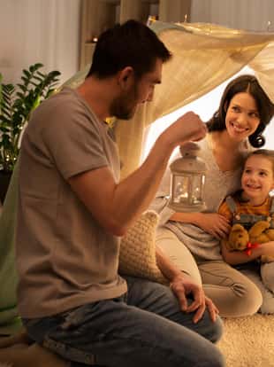 family, hygge and people concept - happy mother, father with lantern and little daughter with teddy bear playing in kids tent at night at home