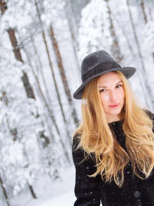 Portrait of a young blonde fashion model walking in the snowy forest, wearing warm black clothing and a hat.