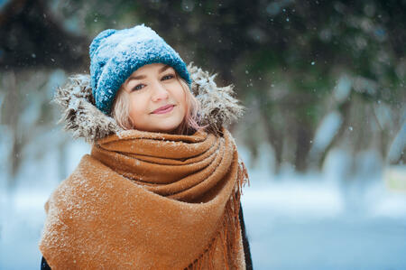 winter portrait of happy young woman walking in snowy forest in warm outfit, knitted hat and oversize scarf. Exploring nature on winter and Christmas vacations concept
