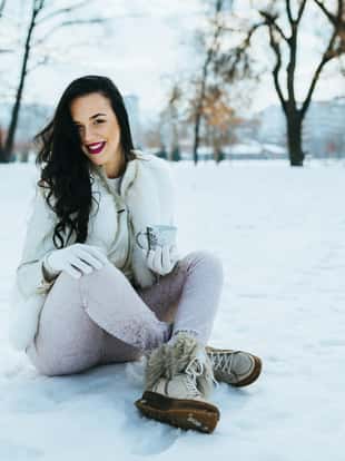 Beautiful young woman sitting on a snowy winter day in the city park, holding a cup and drinking hot beverage