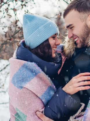Young couple in love walking and laughing in winter forest. People have fun under falling snow covered with blanket to warm up