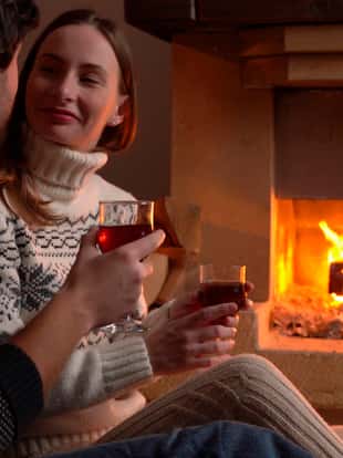 Young romantic couple sitting in front of fireplace at home, drinking red wine