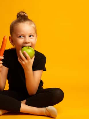 little dancer with apple during break Full body little girl in leotard, nibbling a green apple and holding a carrot while sitting against yellow background during break in training. Healthy food