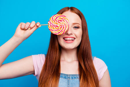 Close up photo beautiful amazing she her lady laugh laughter hiding eye big lolly pop spiral twist candy long wooden stick sweets lover wear casual jeans denim overalls clothes isolated blue background.